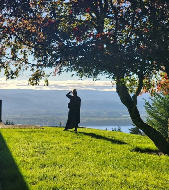 A shot of Debbie from behind wearing a long black dress with her hand shielding her eyes from the sun, standing on a grass lawn looking out over a beautiful landscape below of forests and a river with a large tree framing the photo. She is standing in Morgan Berry's front yard on the top of Elephant Mountain.