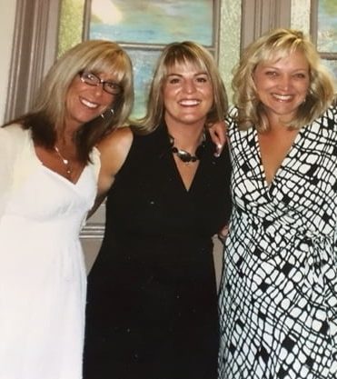 An image of Leslie, Debbie, and Tina all smiling brightly for the camera. Leslie is wearing dark glasses, has shoulder-length blonde hair and wearing a white dress, Debbie is wearing a black dress with a black chunky necklace and Tina is wearing a black and white dress with shoulder-length blonde hair. This image was taken at the wedding where Debbie was ordained to marry her friends, Matt and Kristen.