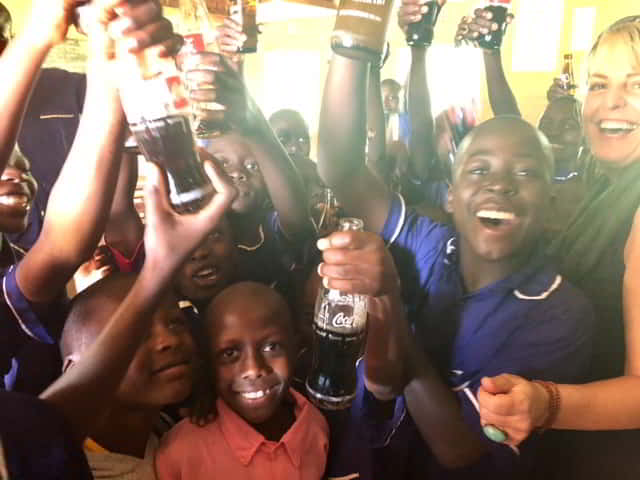 A large group of young Kenyan students and Debbie Ethell off to the side laughing and smiling for the camera as everyone holds bottles of Coca-Cola in the air.