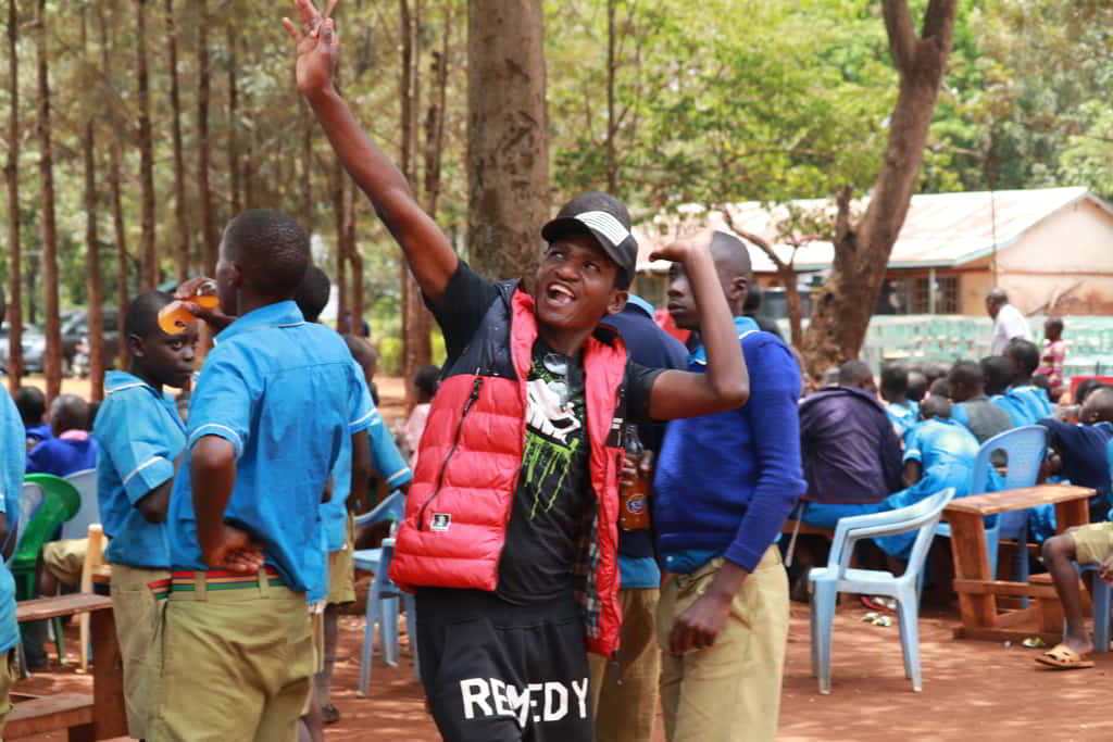 Ibrahim smiling with his hands raised in celebration surrounded by students and newly built desks in the background.