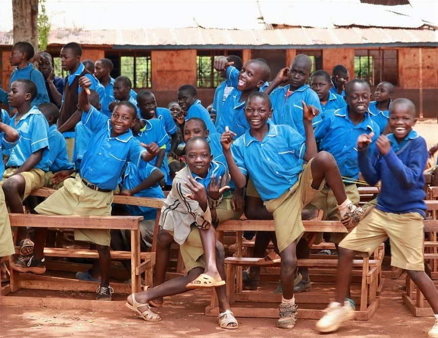 Group of Kenyan schoolboys in uniform dancing and celebrating with newly built desks in the background.