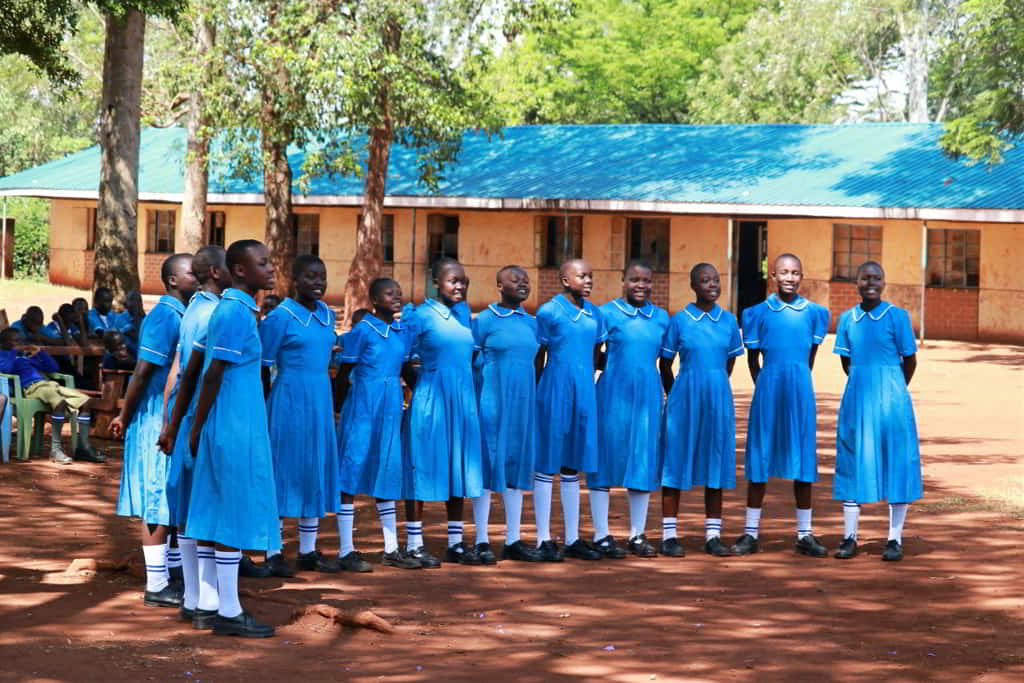 Group of Kenyan schoolgirls in bright blue uniforms arranged in a semicircle signing during a ceremony for a desk donation campaign.